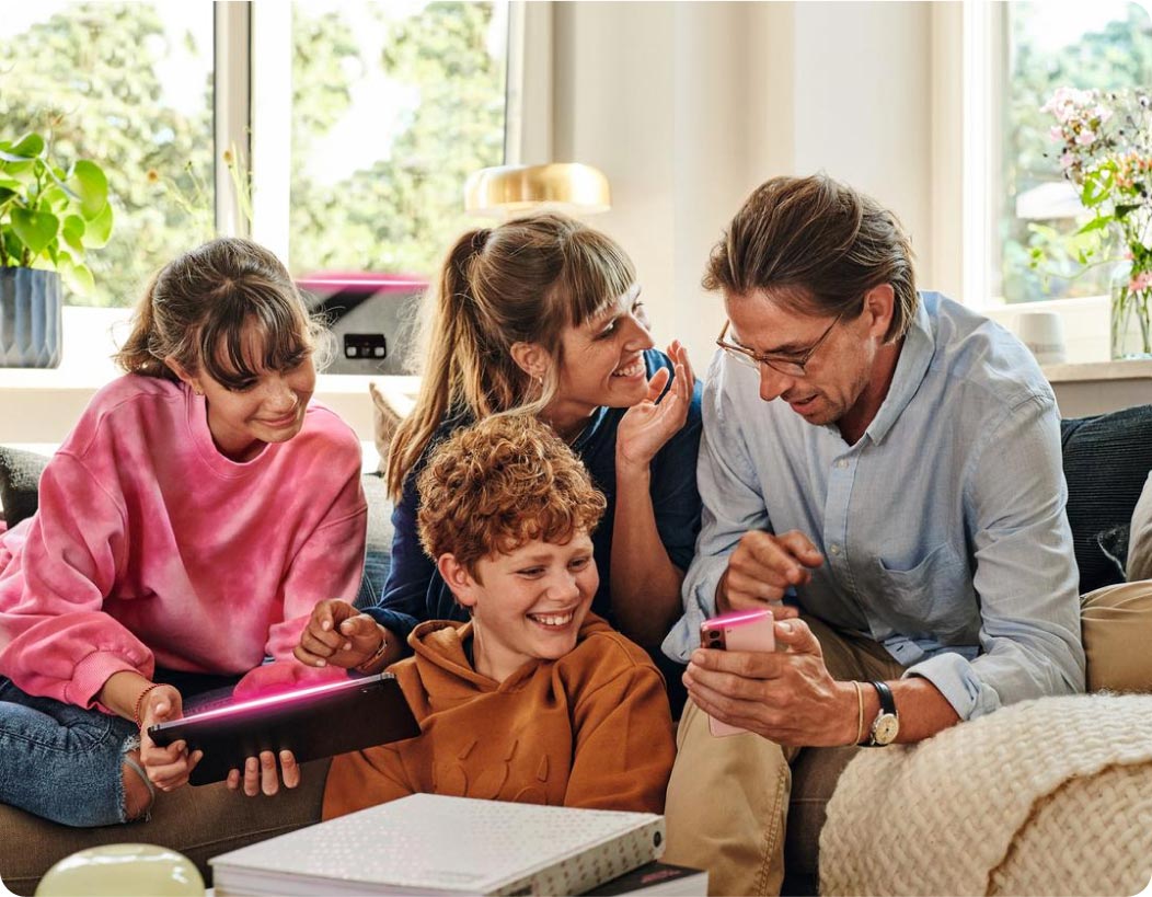 Zwei Personen einer fröhlichen Familie halten ein Tablet und Smartphone in den Händen. Im Hinterground ist ein Glasfaser Router. Alle Geräte leuchten in Magenta.